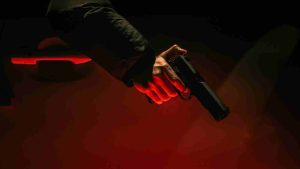 Man holding a gun with his right hand pointing at the ground against red backdrop