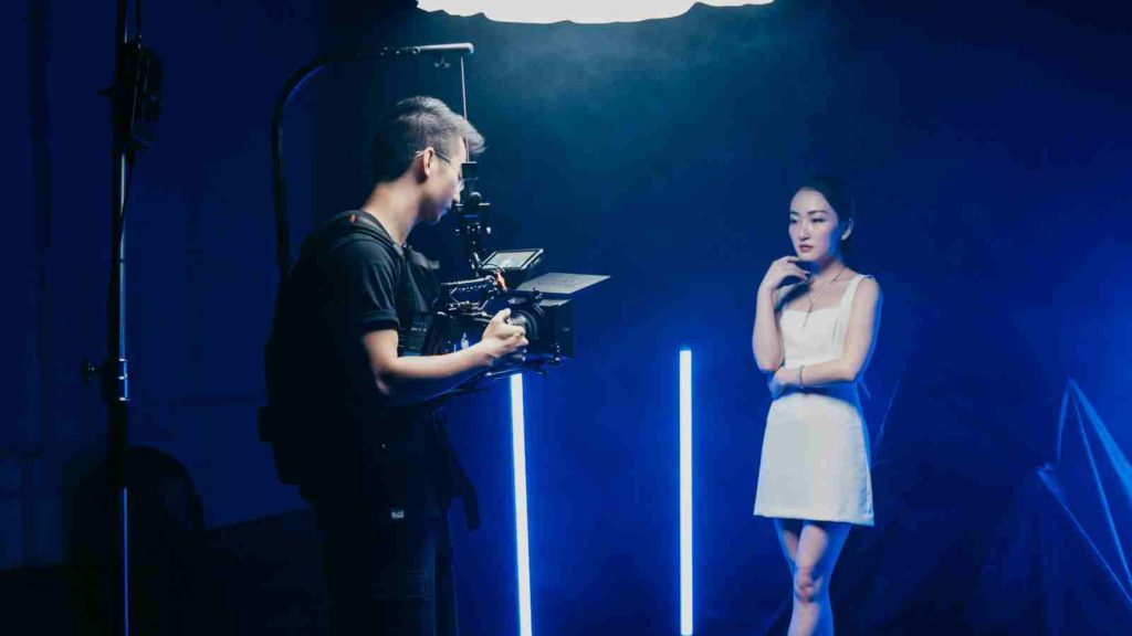 Asian actress in white dress standing under bright light being filmed by cameraman