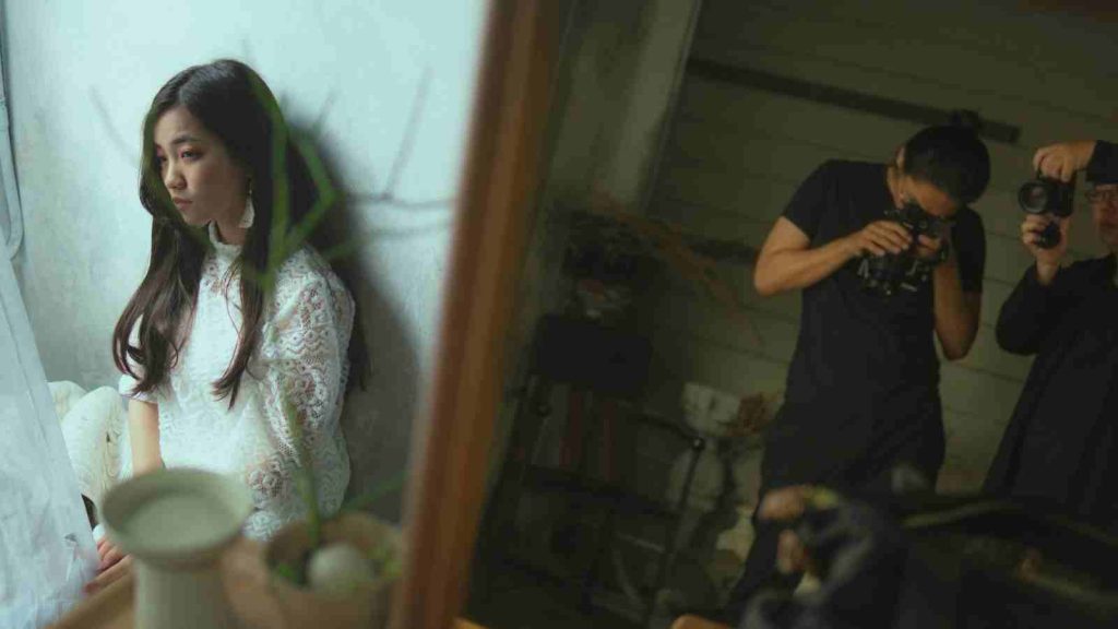 Asian actress in white dress waits in the room while cameraman are outside adjusting filming equipment