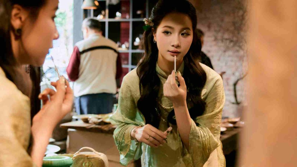 A Vietnamese girl in a turquoise ao dai dress putting on lip gloss in the mirror in a cafe