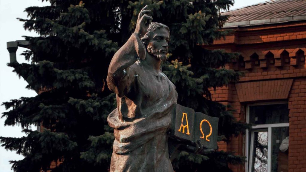 A bronze statue depicting a historical figure holding a book with Alpha Omega symbol, outdoors near a brick building.